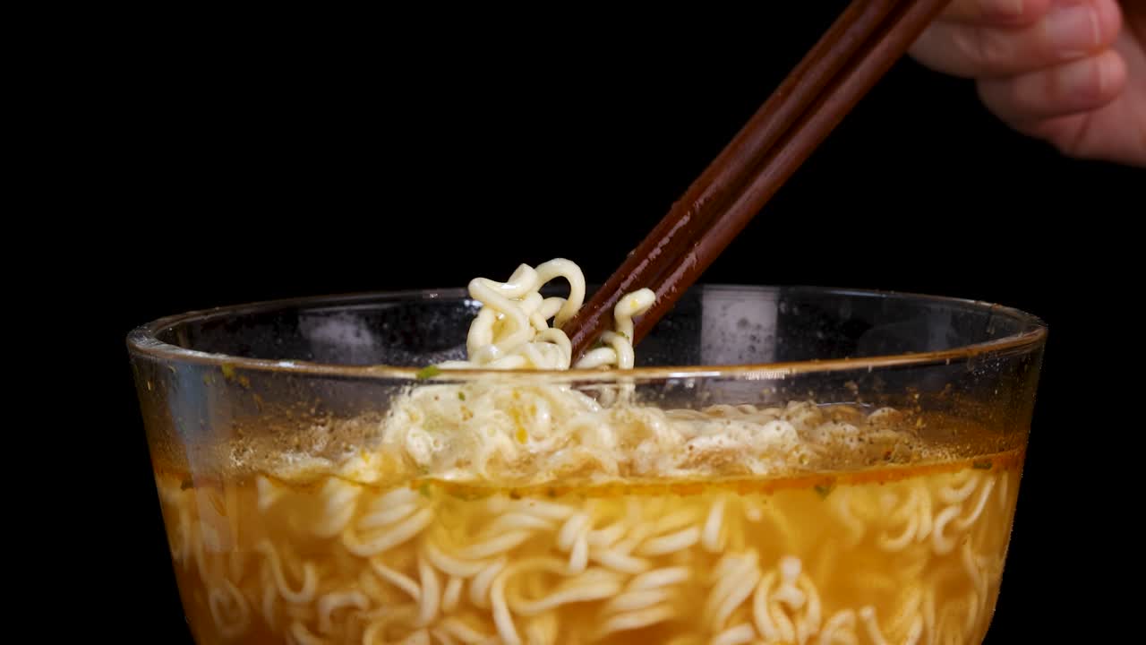 Hand uses chopsticks to lift steaming instant noodles from a clear glass bowl filled with broth, set against a black background with even lighting