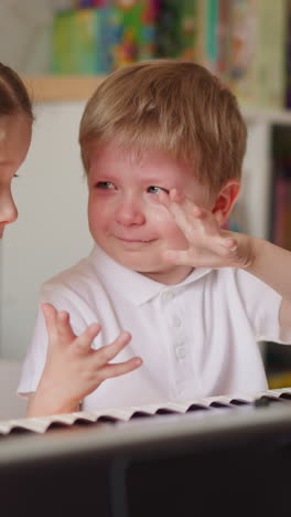 little boy smiles through tears and shows hands to sister at piano in play room. girl supports pensive toddler kid tired of music chords learning