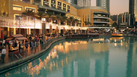 people walking through the streets under the burj khalifa, dubai, during the starting period of corona virus. slow motion pan of walking street and artificial lake under the worlds largest building.