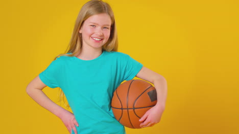 studio shot of young girl holding basketball under arm against yellow background