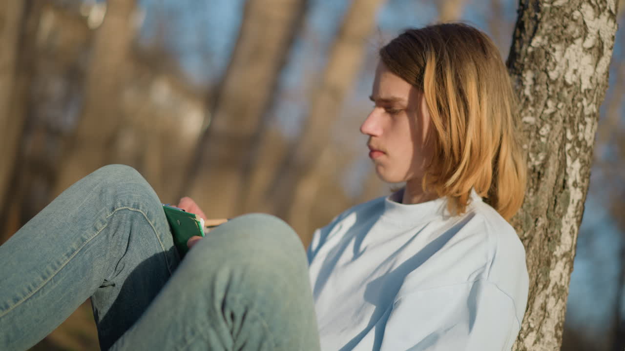 Adolescente junto a un abedul, joven apoyado cómodamente en un esbelto abedul blanco; una persona adolescente se toma un momento de reflexión mientras se apoya en un elegante abedul blanco.