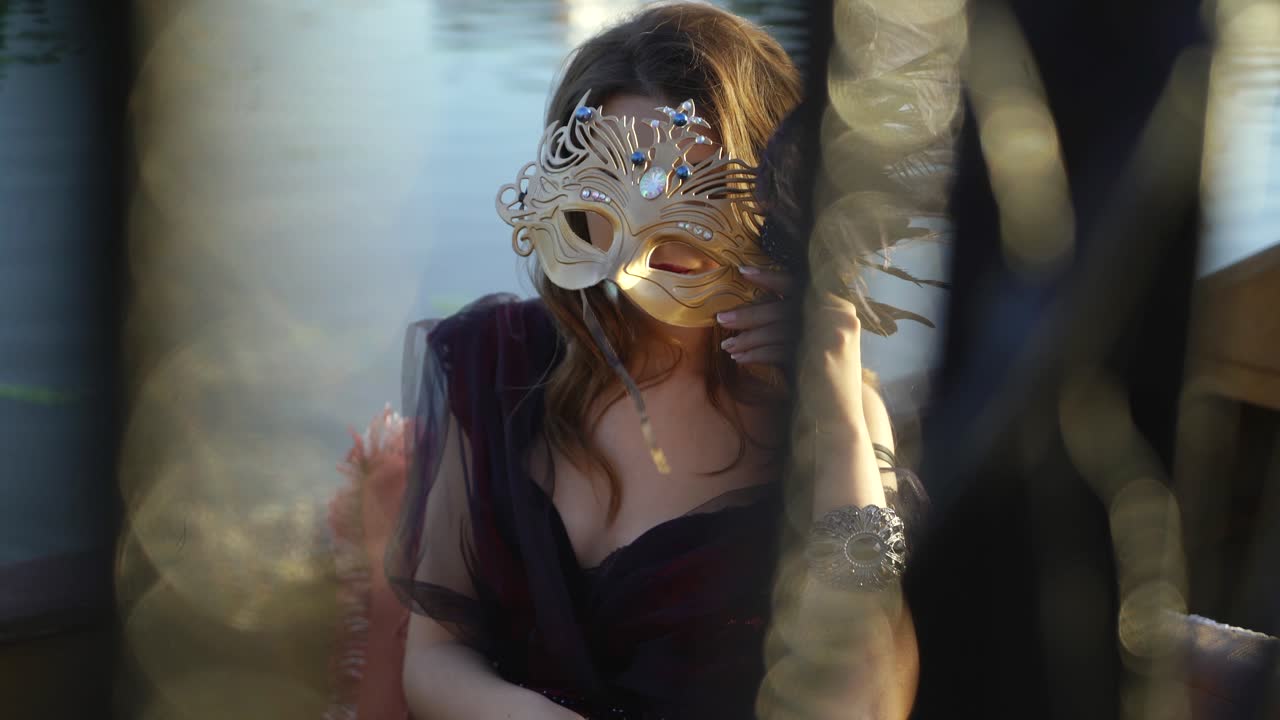 Sexy woman closes her face with the carnival mask on the blurred background. Beautiful young lady in holiday make up looking at camera. Close-up
