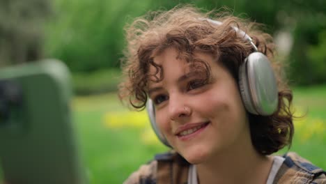 Close-up-a-girl-with-curly-hair-in-wireless-headphones-turns-on-tracks-on-a-green-phone-and-listens-to-music-in-the-park