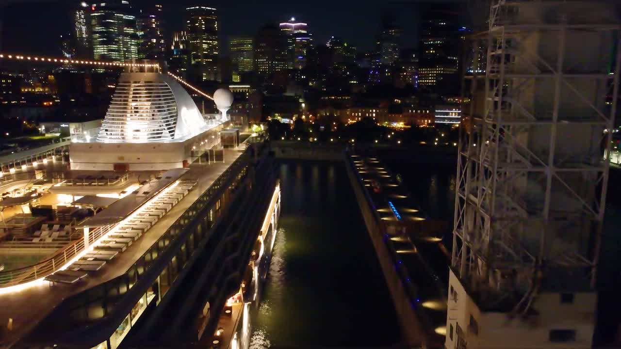 Drone night flight tilts up to reveal illuminated Regent Seven Seas Grandeur cruise ship, city lights, skyline, harbor - Montreal, Canada