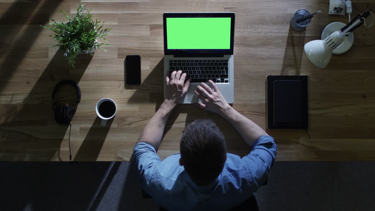 vista superior de un estudiante masculino trabajando en su computadora de escritorio con una maqueta de pantalla verde por la noche. su mesa está iluminada por una luz azul fría desde afuera.