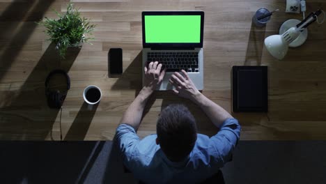 vista superior de un estudiante masculino trabajando en su computadora de escritorio con una maqueta de pantalla verde por la noche. su mesa está iluminada por una luz azul fría desde afuera.