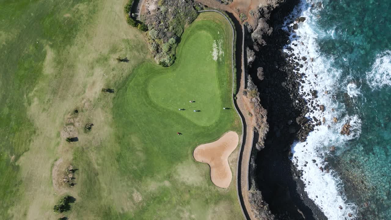Drone top down bird's eye view of golfers on putting green as clear ocean water waves crash on rocks
