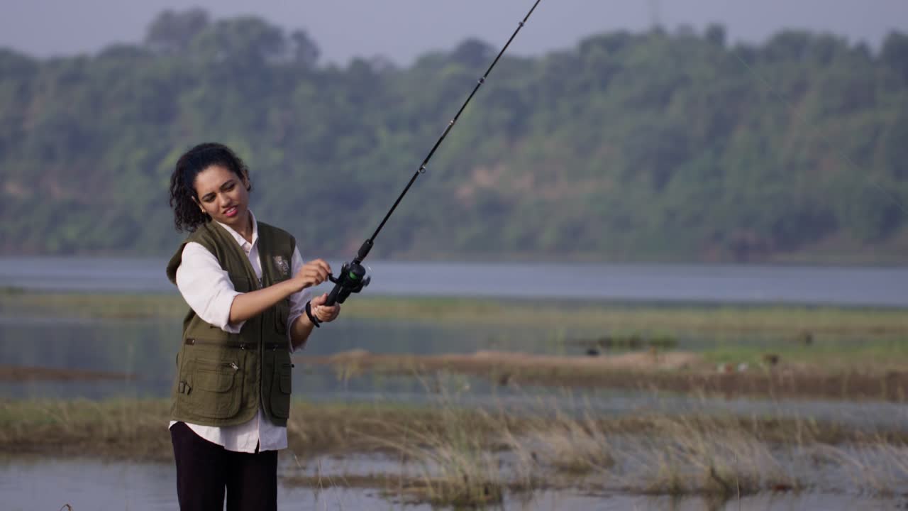 Seorang gadis Asia Selatan mengenakan rompi memancing memegang pancingnya di dekat danau yang tenang, tersenyum lembut dalam cahaya alami.