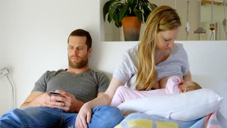 front view of father using mobile phone while mother breastfeeding to baby on bed at home 4k