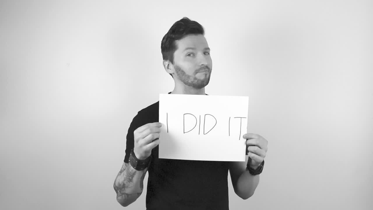 Grayscale footage of a tattooed man with short hair and trimmed beard lifting up a white big paper sign that says I Did It in capital letters while the man is confident and is smiling.