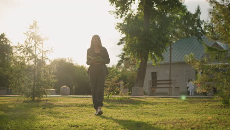 lady in grey clothing reading a book while walking on grassy field outdoors during golden hour, sunlight illuminates the surrounding, with two persons walking near a building in the background