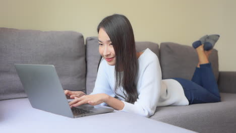 close-up of an attractive young woman in jeans and a sweatshirt working on her laptop while lying on her stomach on the sofa