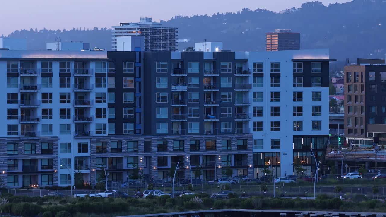 Vista aérea ascendente de drone de edifícios residenciais modernos à beira-mar em Oakland, Califórnia, com colinas distantes visíveis ao fundo enquanto as luzes da noite se acendem