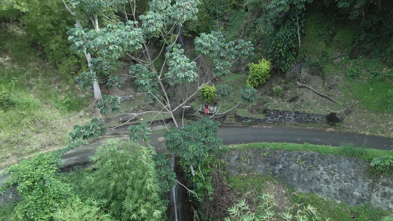 vista aérea de una mujer joven sentada en un banco en un bosque con una cascada que cae por la montaña