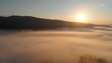 aerial shot of a golden sunrise shining on clouds covering mountain peak with hills on the horizon