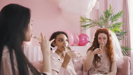 Side-View-Of-Three-Multiethnic-Friends-Talking-And-Drinking-Champagne-Sitting-On-Sofa-Celebrating-Bachelorette-Party