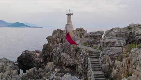 Isla-De-Kalamota,-Mar-Adriático,-Croacia---Una-Mujer-Vestida-Con-Un-Vestido-Carmesí-Saborea-La-Puesta-De-Sol-Junto-Al-Faro---Disparo-De-Drone-En-órbita