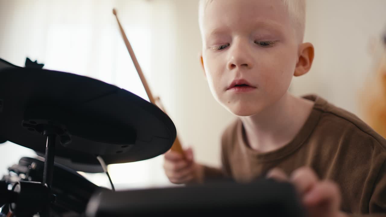 Close-up shot from below of a happy blond albino boy with white hair setting up an electronic drum kit before starting to play it with wooden sticks in his room, developing his talent and hobby