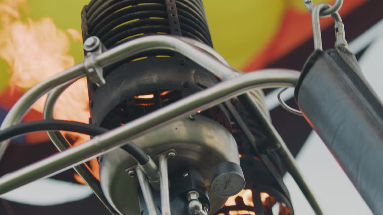 Close up of metal burner on hot air balloon with visible fire glowing inside heating chamber, surrounded by steel pipes and gauges, viewed from below during ignition process under bright daylight