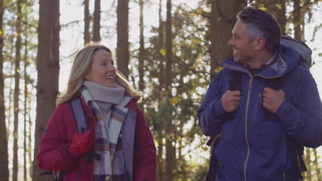 Front-view-of-mature-couple-with-backpacks-walking-through-fall-or-winter-countryside-against-flaring-sun--shot-in-slow-motion