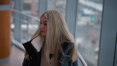 young lady gazing through glass window of building as she looks back contemplatively, her hair cascading down her jacket with urban city view visible through glass in the background