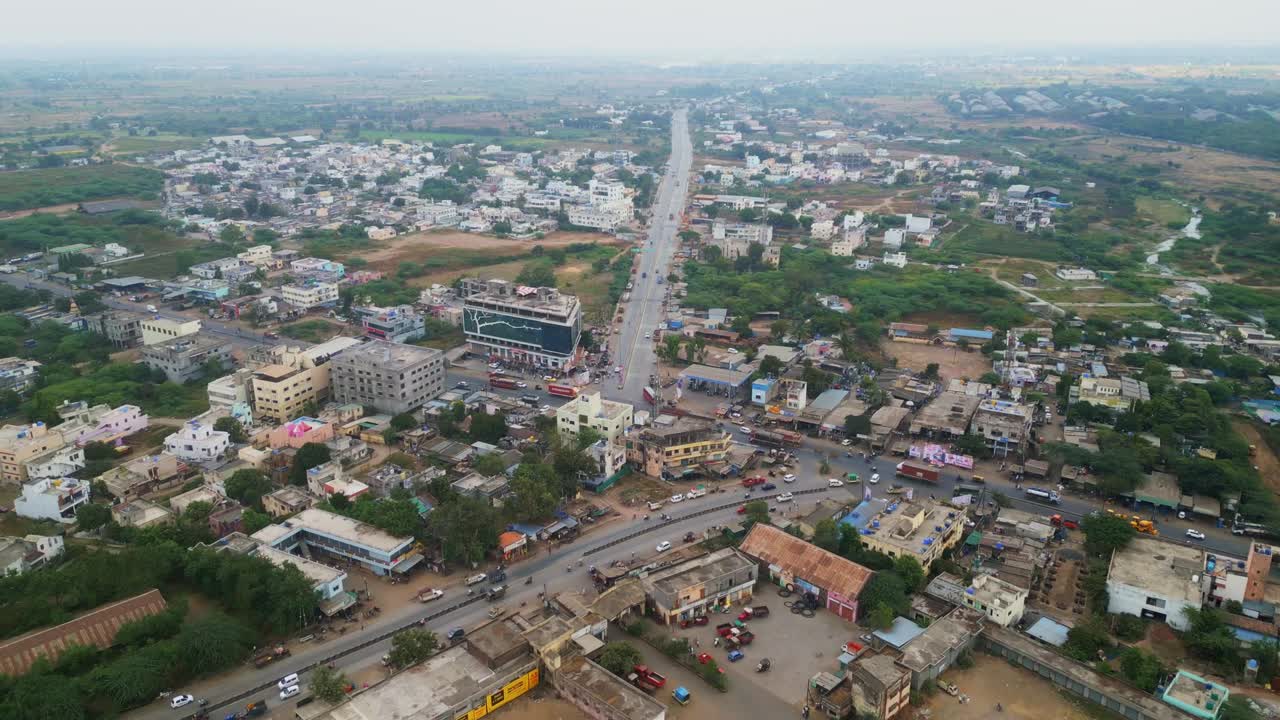 A cinematic drone view captures vehicles moving along a main road in Manmad city, Nashik, Maharashtra. Buildings line both sides under a bright sunny sky, showcasing vibrant urban life in motion 4K