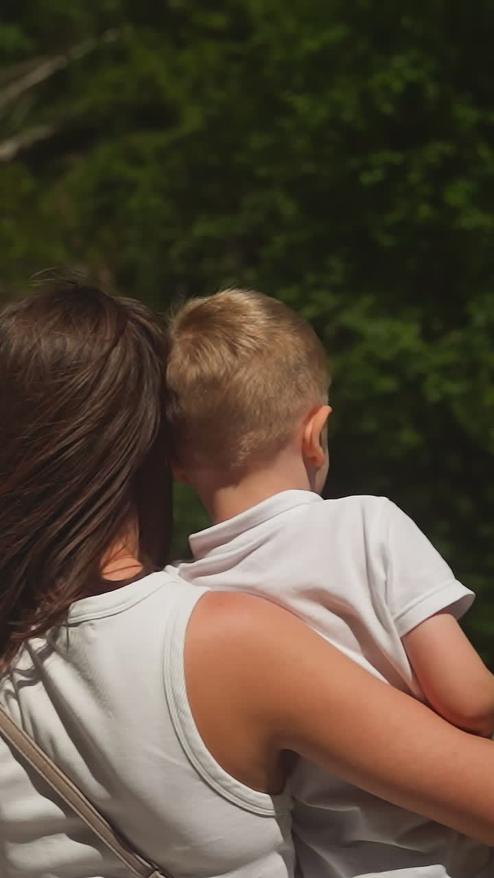 Happy mother with little boy enjoys wonderful waterfall on cliff in summer park. Woman and toddler son look at mountain river cascade in forest