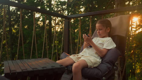 little girl enjoys carefree childhood amidst nature sitting in chair on terrace. girl watches movie on smartphone while sitting in armchair at calm sunset