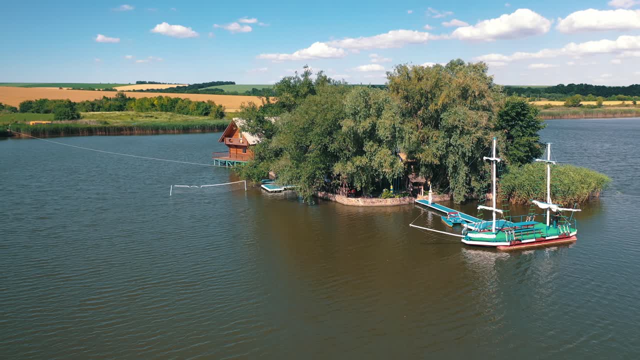 Drone flying over the river to island at daytime in summer. Small island with green trees and an old boat outdoors. Aerial view. Camera moves forward.