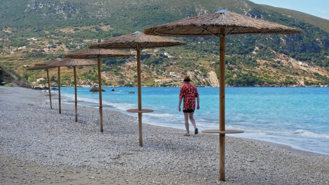 A Man In Summer Beach Outfit Walking Along The Shoreline Pebbled Beach Of Agia Kiriaki, Zola In Kefalonia, Greece