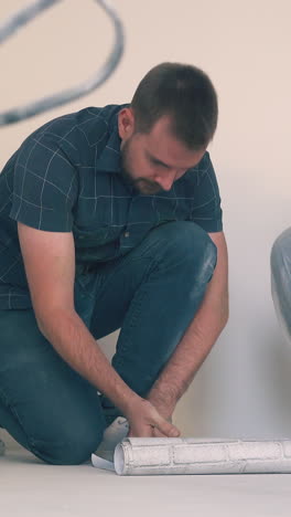 dolly cam: happy young family of bearded man and blonde girl unrolls sheet of light wallpaper on floor in empty room