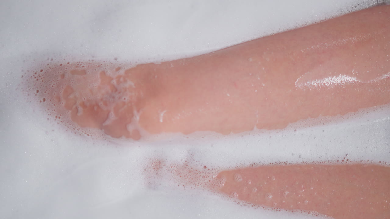 mujer con las piernas desnudas en agua espumosa en la bañera. dama delgada toma un baño cálido acogedor con espuma de jabón en casa. mujer elegante se relaja sentada en un baño caliente
