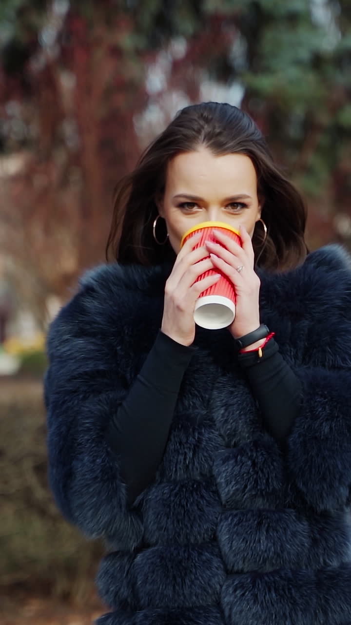 Beautiful girl drinks coffee outdoors. Portrait of a fashionable woman in fur coat with hot drink looking on camera. Stylish model in the park. Vertical video