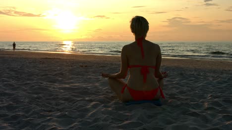 cute slim woman in bikini during vacation at the seaside resort sits in a lotus flower position on a sandy beach and meditates while the sun hides over the horizon