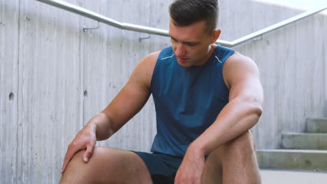 Young-Man-Massaging-Injured-Leg-Sitting-on-Stairs.fitness,-sport-and-health-concept-young-man-massaging-injured-leg-or-knee-sitting-on-stairs