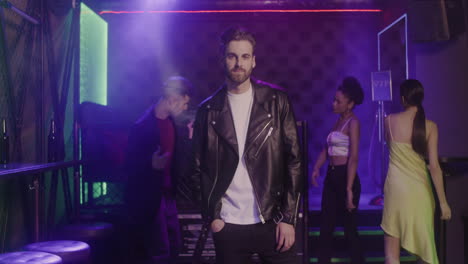 Handsome-Bearded-Man-In-Leather-Jacket-Smiling-At-Camera-At-Disco-While-His-Friends-Dancing-Behind-Him