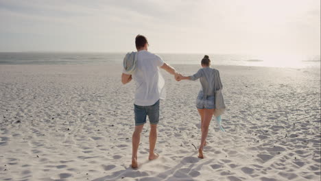 Young-couple-holding-hands-walking-towards-sunset-on-empty-beach
