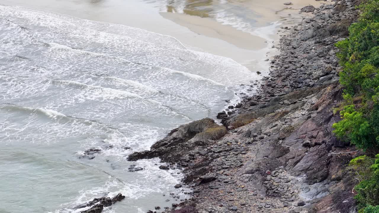 Aerial footage captures a rocky shoreline transitioning to a serene tropical beach with lush greenery in Port Douglas