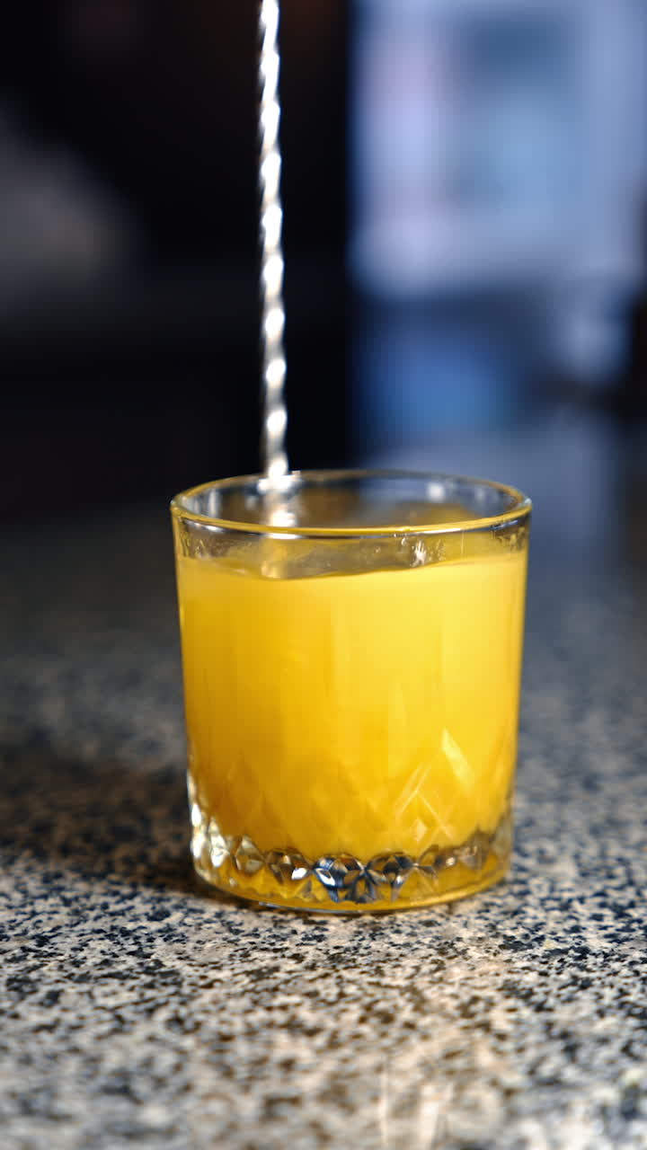 Yellow drink is mixed in the glass with an ice cube. Long spoon stirring the cocktail. Close up. Vertical video.
