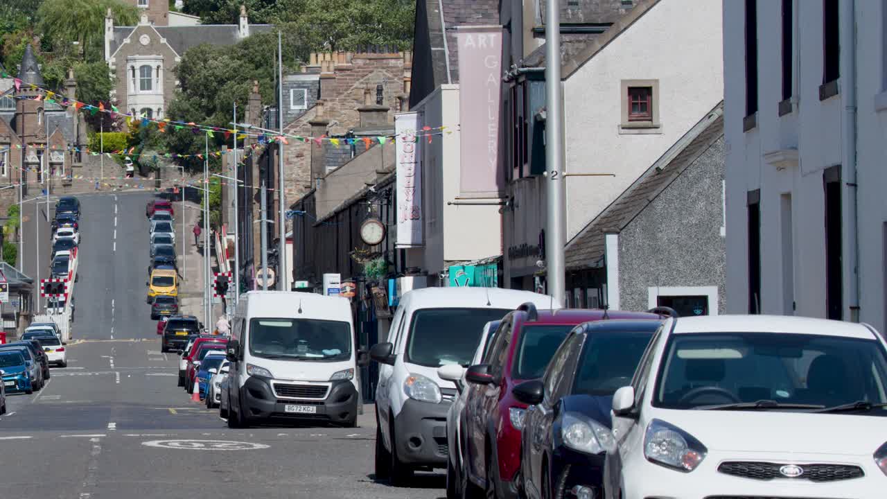 Stationary camera captures parked cars, pedestrians, and buildings on a sunlit, sloping urban street