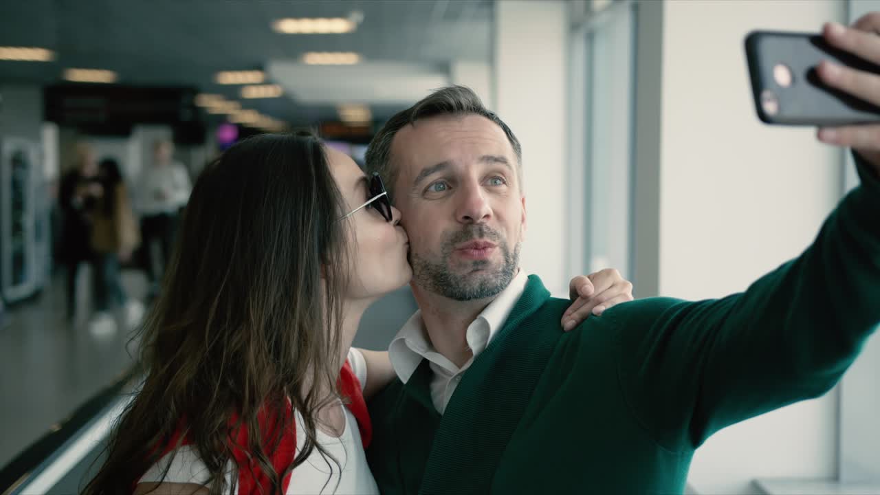 Young beautiful couple in the hall of airport. Brunette smiling woman and man taking selfie photo on smartphone near the window. Cinematic shot on RED camera. 2 models.