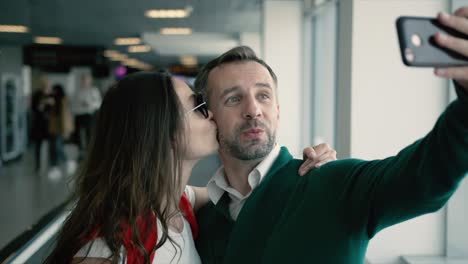 young beautiful couple in the hall of airport. brunette smiling woman and man taking selfie photo on smartphone near the window. cinematic shot on red camera. 2 models.