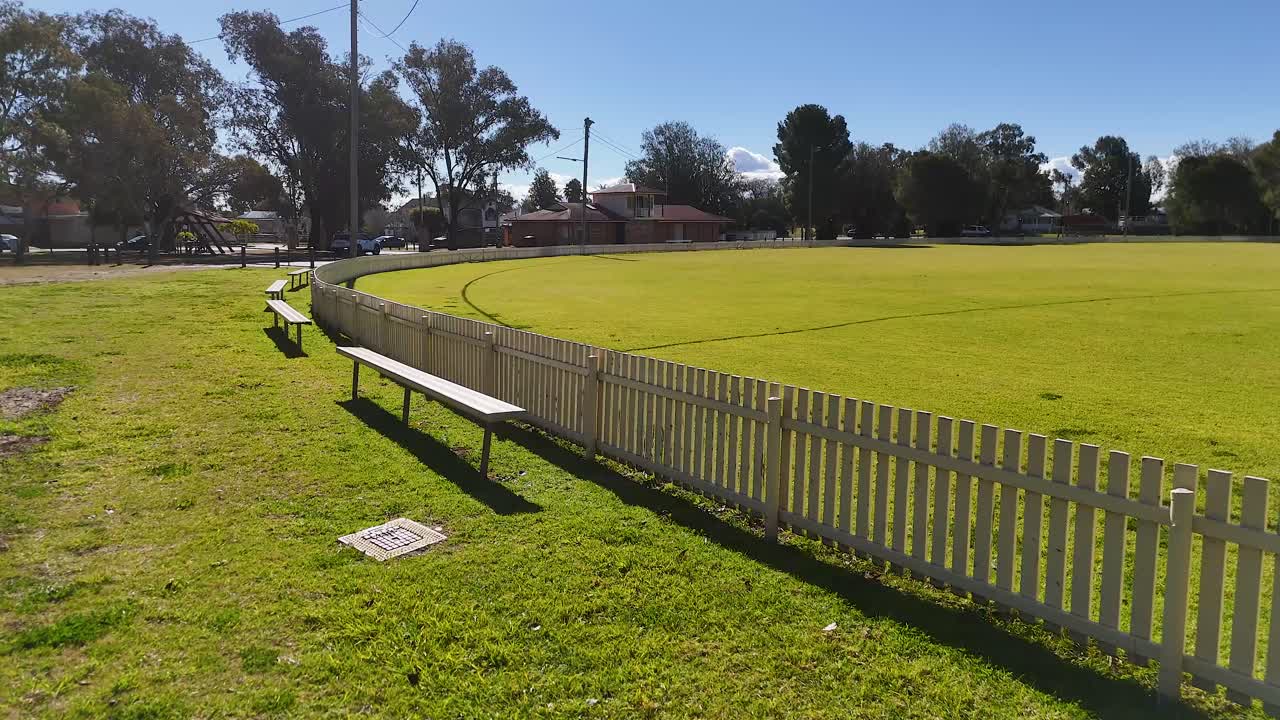 Camera smoothly pans along a white fence bordering a green sports field under clear blue sky, revealing benches, goalposts, and open grassy space