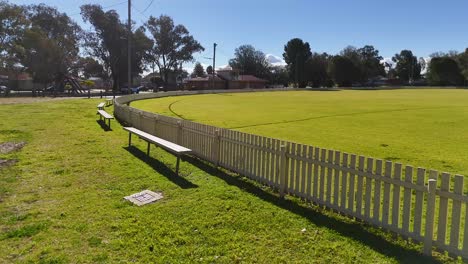 camera smoothly pans along a white fence bordering a green sports field under clear blue sky, revealing benches, goalposts, and open grassy space