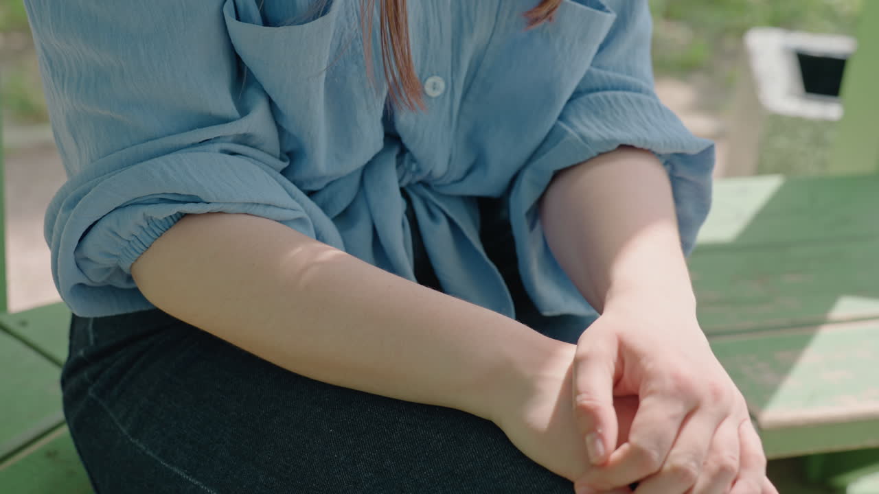 primer plano de una mujer caucásica subiéndose las mangas en un banco verde, parque iluminado por el sol, vaqueros y blusa azul holgada, manos ajustando el puño, sombras suaves, ritmo relajado, detalle íntimo