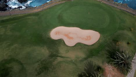 Revealing-Shot-of-Sand-Bunker-on-Green-Golf-Resort-Right-By-Rocky-Shore-in-Middle-of-Blue-Ocean