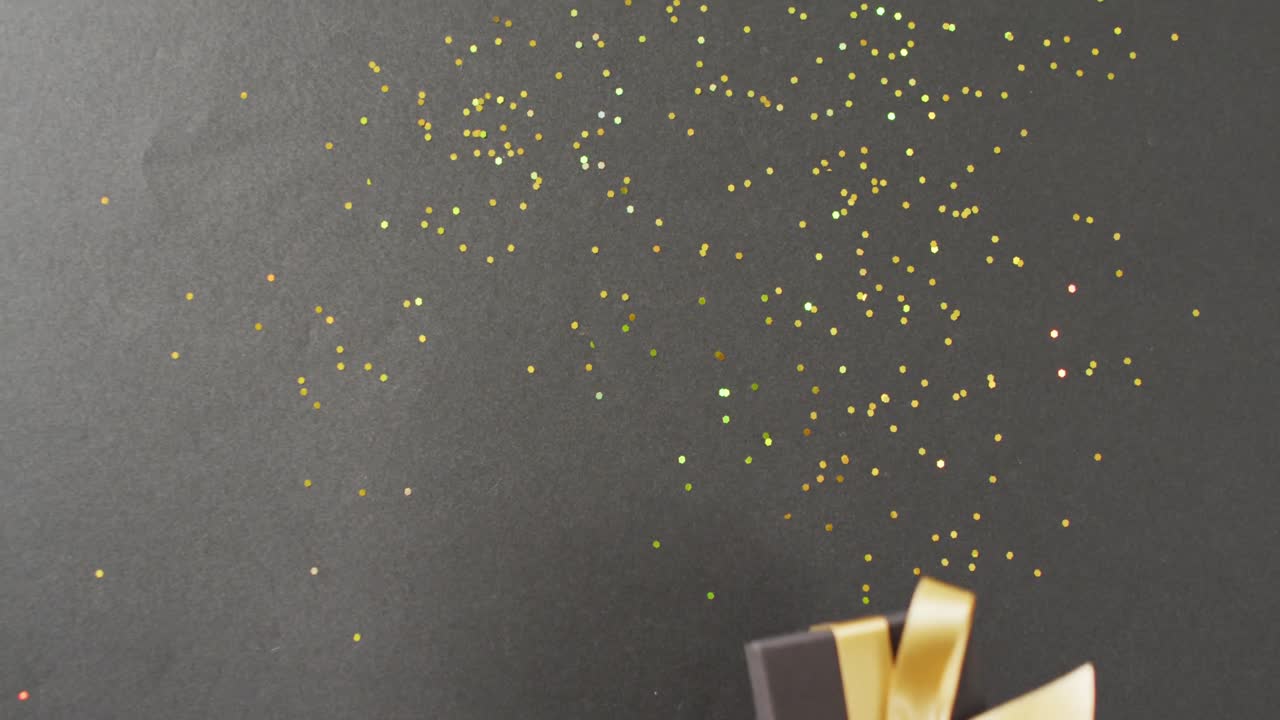 Woman's hand holding black gift box with gold ribbon, over glitter on black with copy space