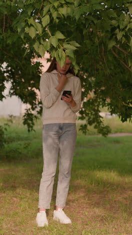 lady in casual attire stands thoughtfully under tree, focused on phone screen with hand touching hood collar, sunlight filters through leaves, with background greenery and glimpse of building