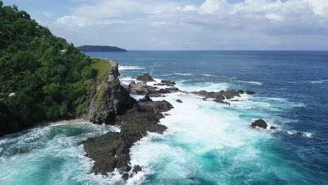 reveal drone shot of waves of indian ocean hitting boulder and hill overgrown by dense of trees beside the beach in sunny condition weather - pengilon hill, indonesia, asia