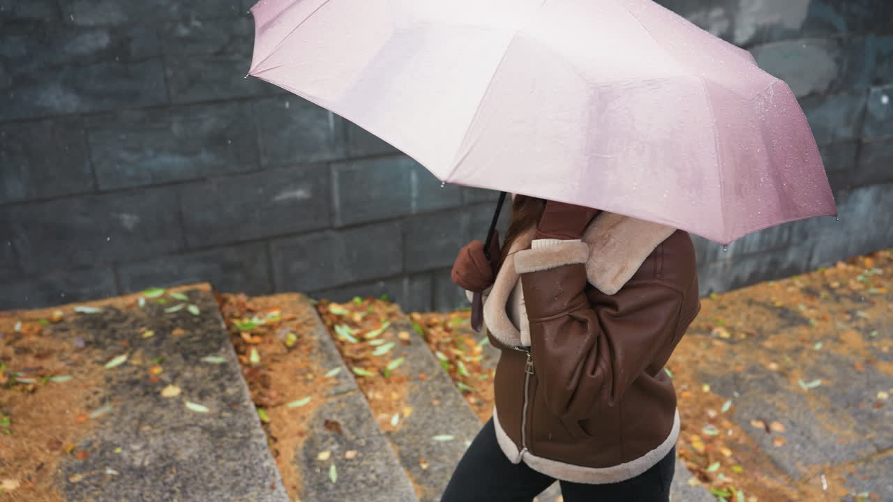 Up shot of young girl smiling on phone call holding umbrella, wearing knit cap, brown shearling jacket, black trousers, walking down stone steps during light snowfall with colorful autumn leaves